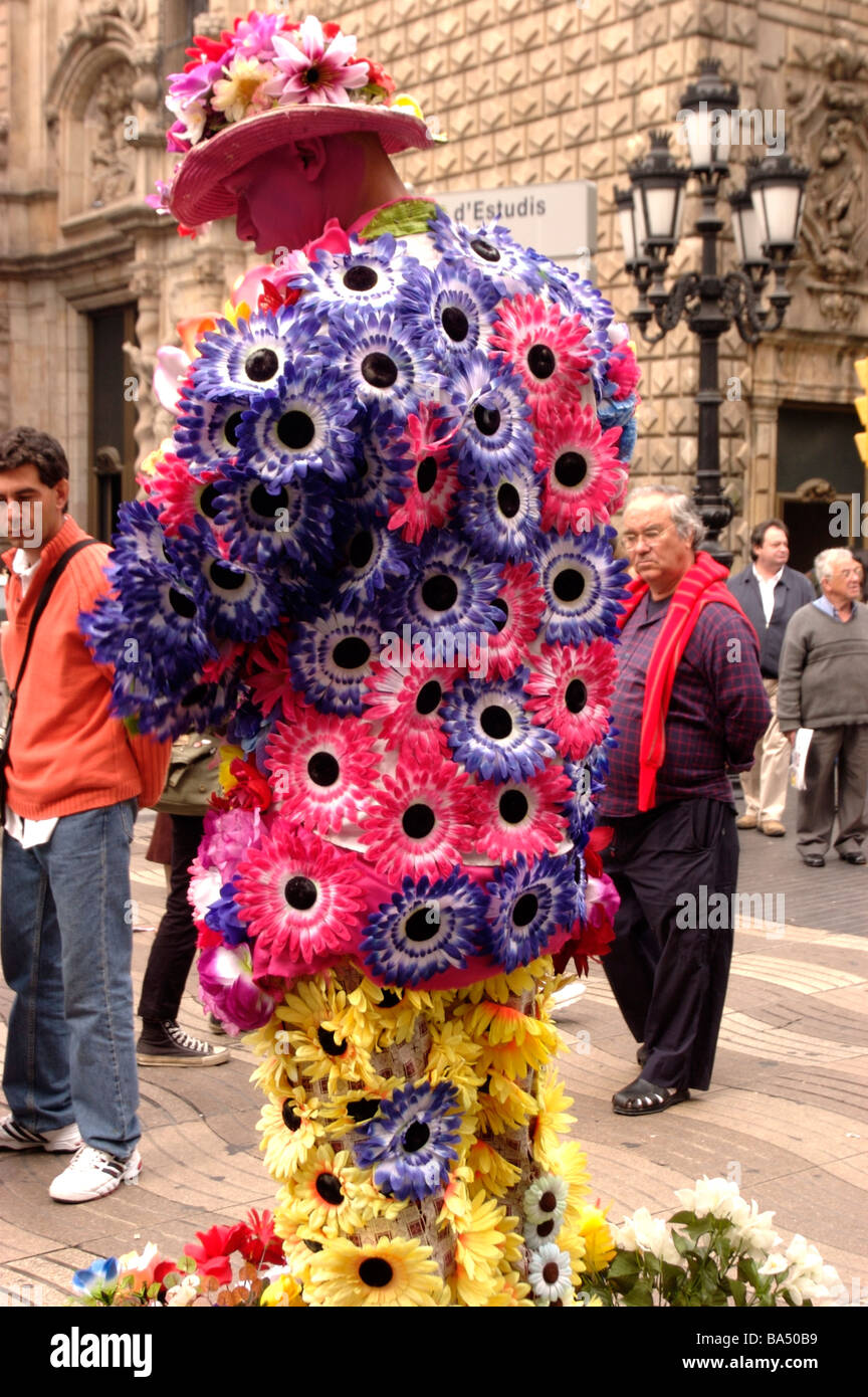 A face painted, street artist dressed in multi colored flowers performs on Los Rambla in Barcelona, Spain with people watching. Stock Photo