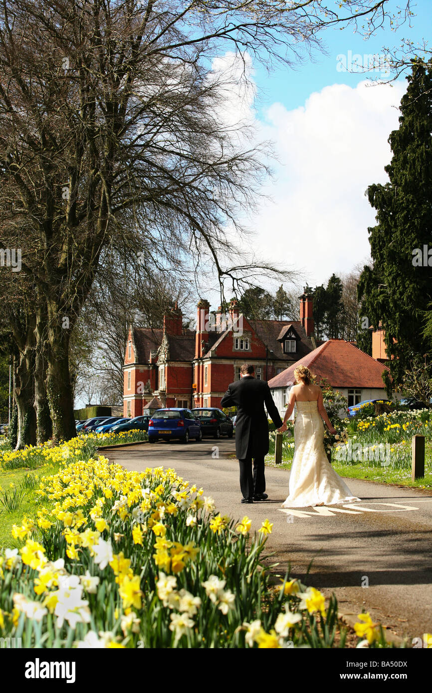 Bride and Groom walk hand in hand on wedding day through spring dafodills in grounds of countryside wedding venue manor house Stock Photo