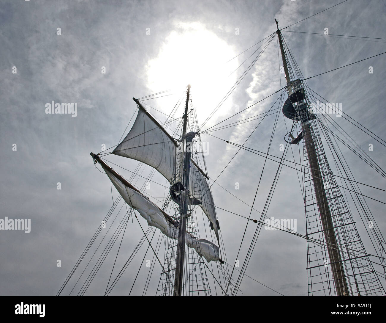 tall-ship tall-ships two of three masts looking upward with bright sun and gray clouds, sail unfurled ropes lines and ladders Stock Photo