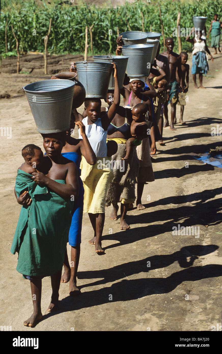A line of women some with babies carry heavy buckets and ceramic pots on their heads after collecting water at a well Malawi Stock Photo