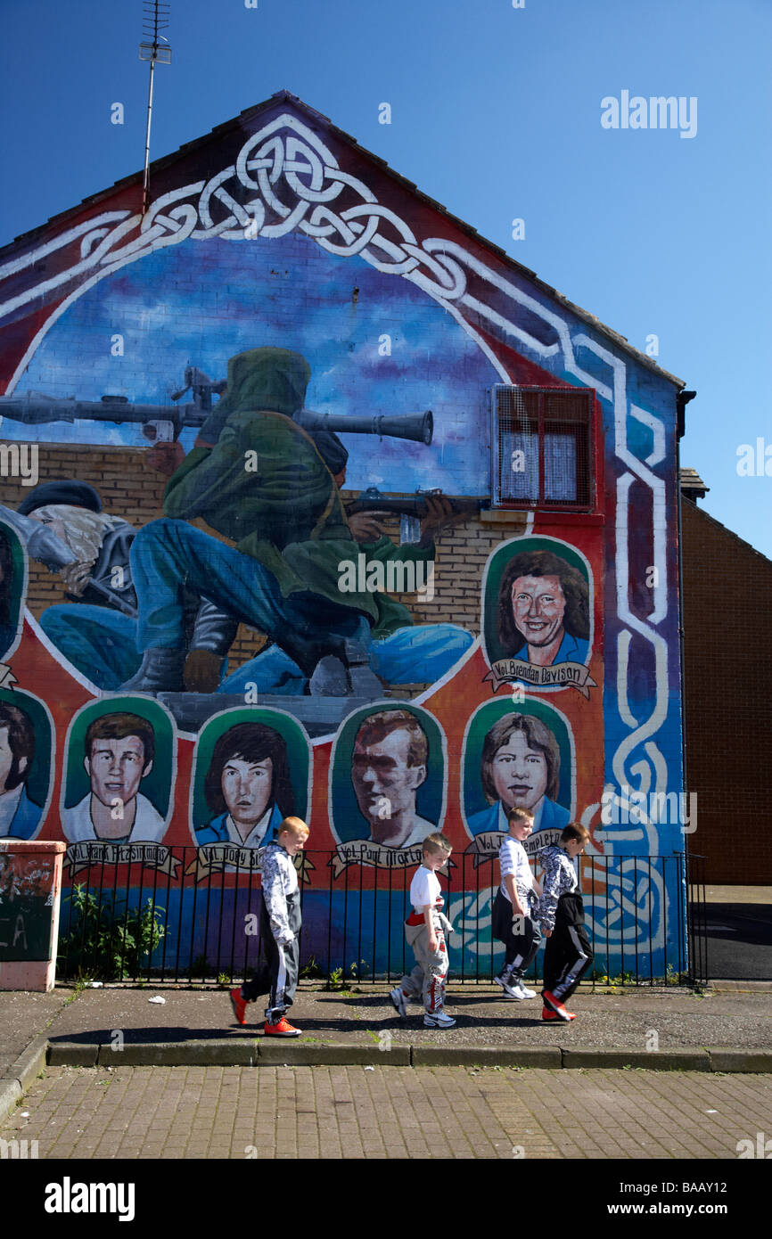 group of young boys walk past an IRA republican mural showing dead volunteers and IRA RPG rocket attack in progress painted Stock Photo