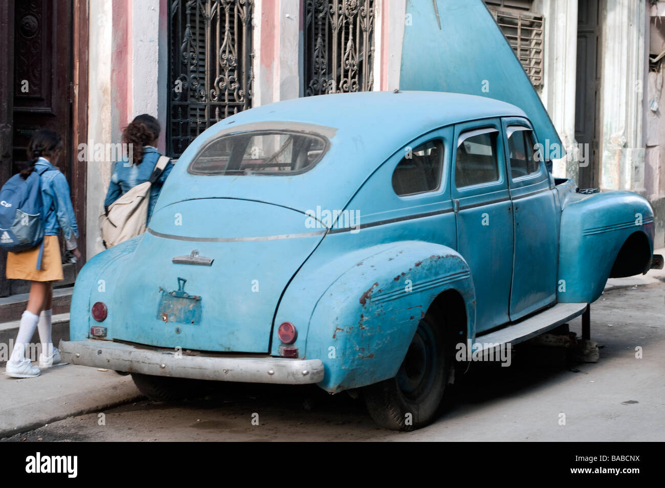 Antique car on blocks on a street in downtown Havana, Cuba with two schoolgirls passing by. Stock Photo