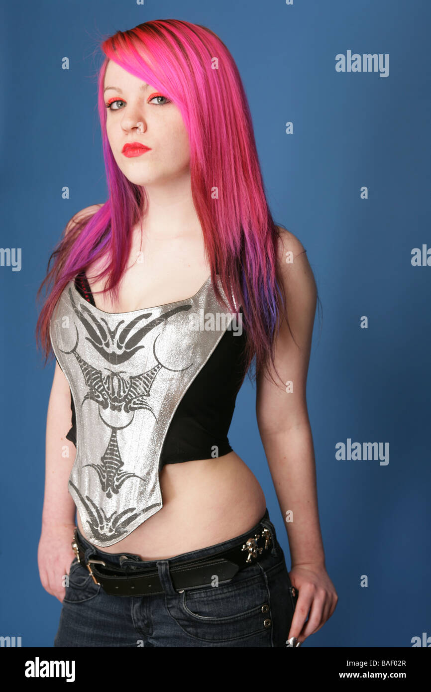 Young teen with bright pink hair red lips and pale skin standing against a blue background looking to camera. Stock Photo