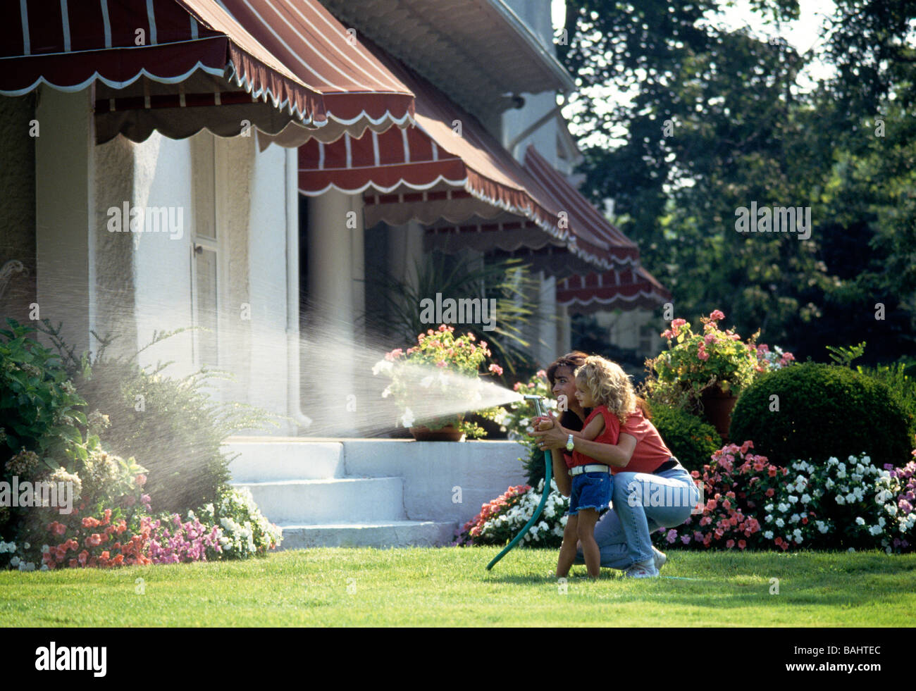 Mother and young daughter using a garden hose to water flower beds on