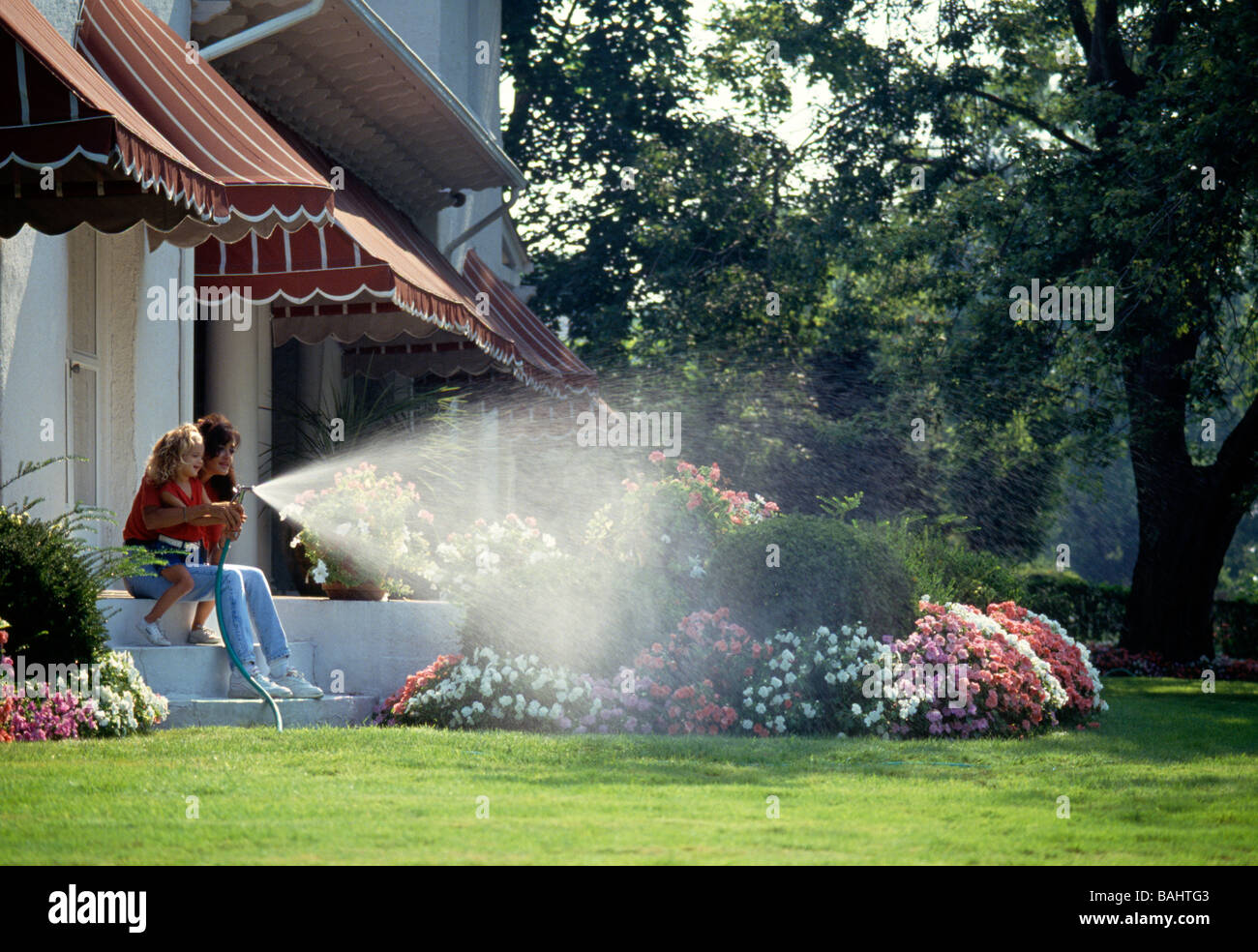 Mother and young daughter using a garden hose to water flower beds on