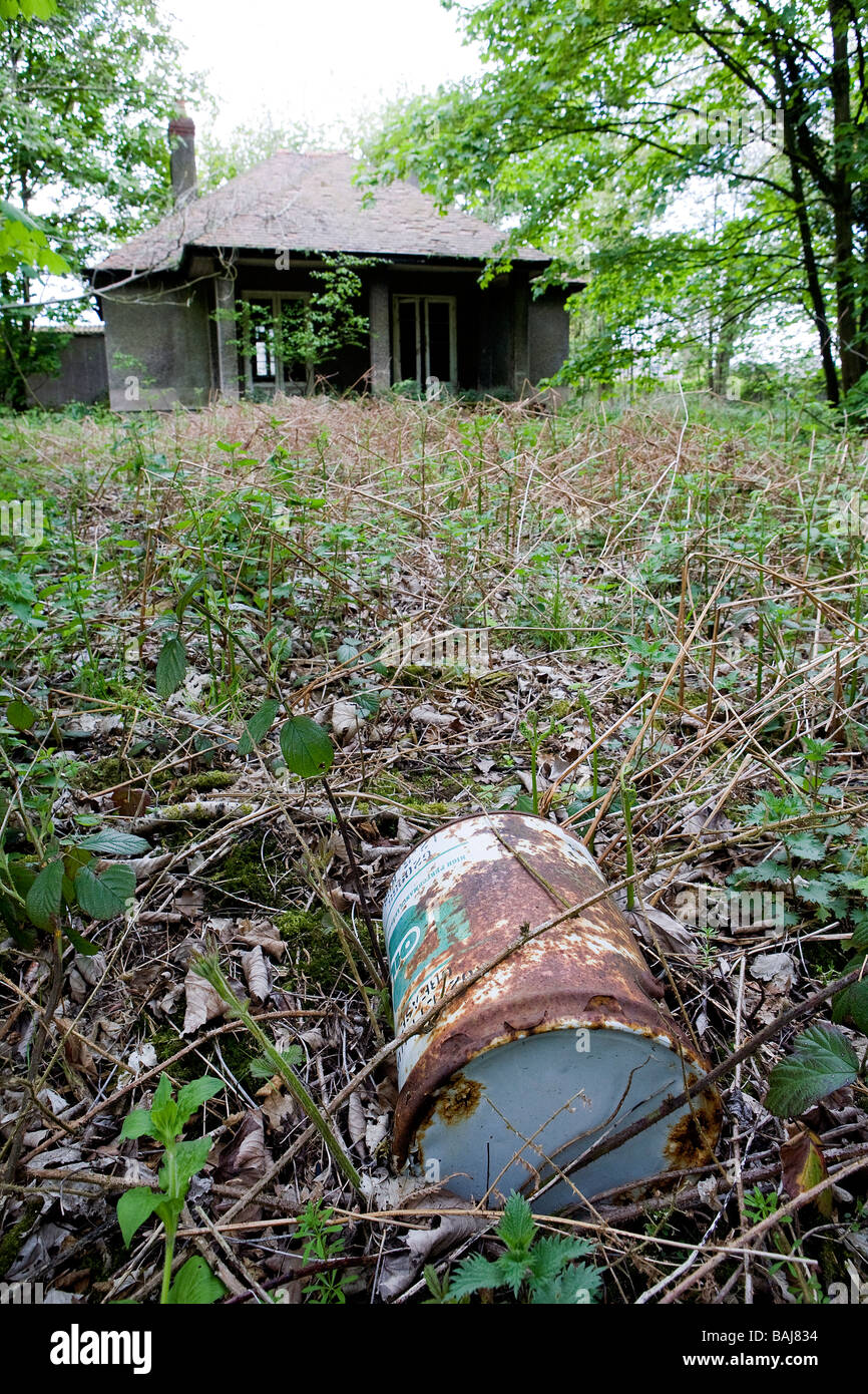 An abandoned cottage lies in disrepair with plants growing through the cracks and an old oil drum in the overgrown garden Stock Photo