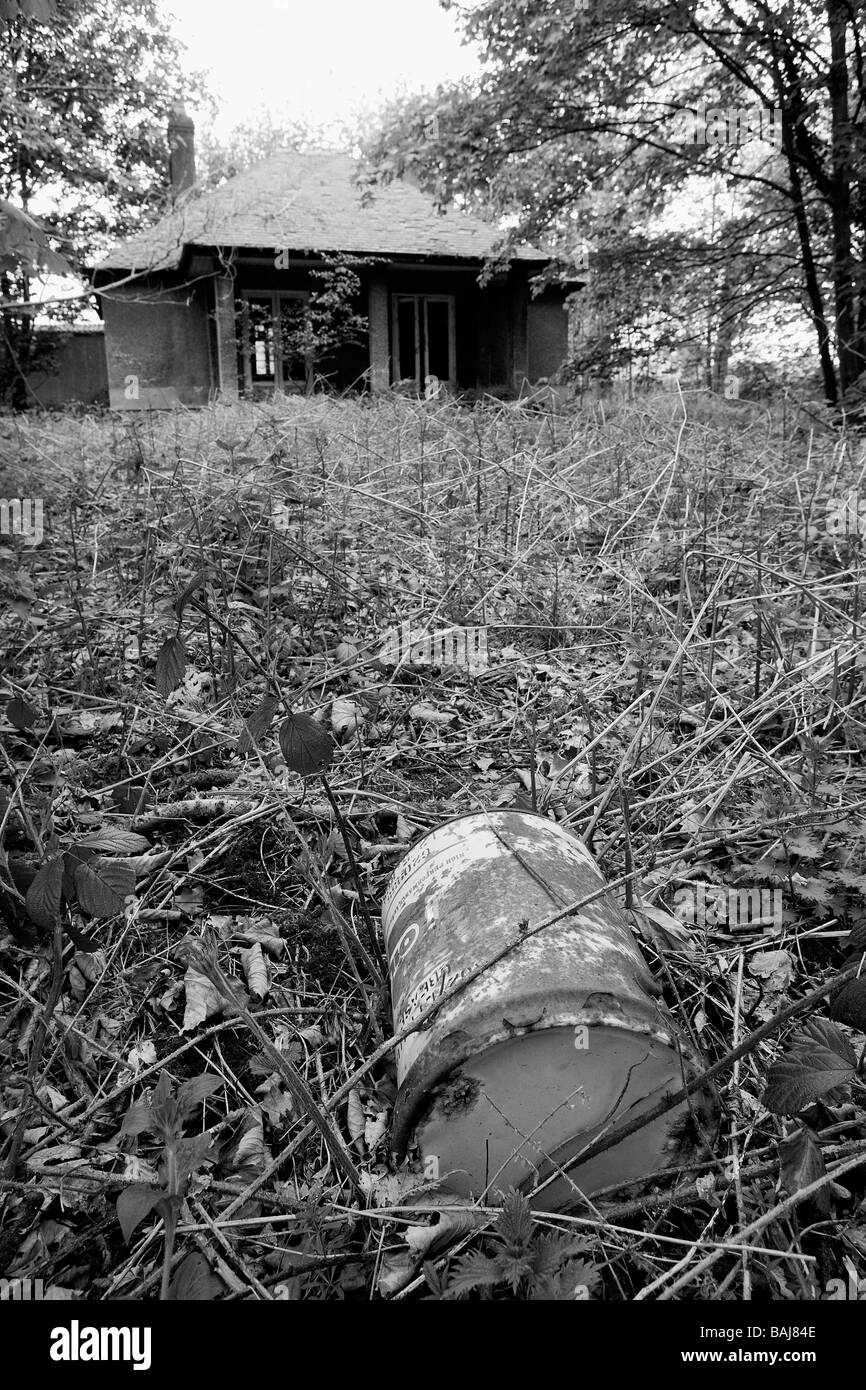 Black + white picture of an abandoned cottage which lies in disrepair and an old oil drum in the overgrown garden Stock Photo