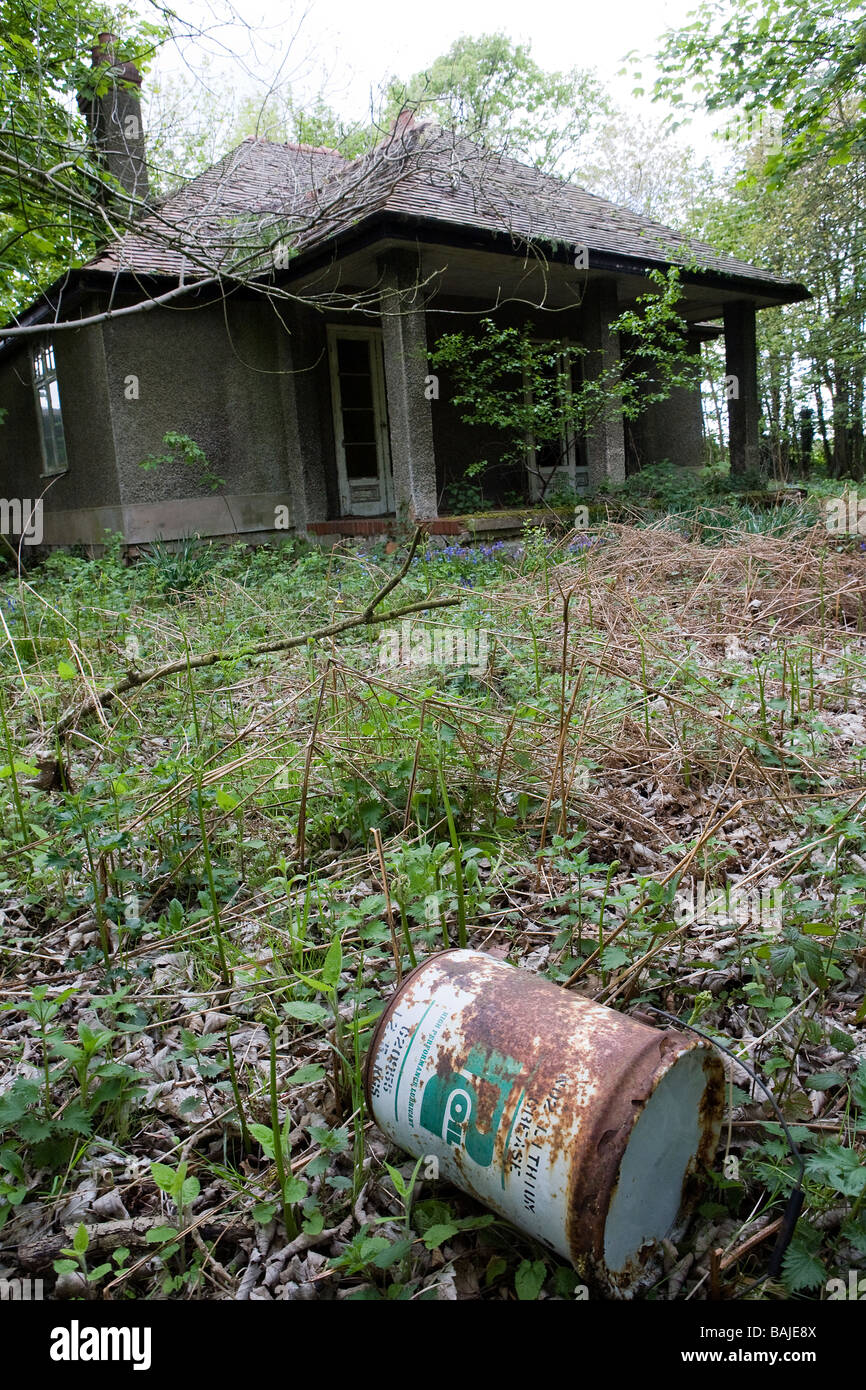 An abandoned cottage lies in disrepair with plants growing through the cracks and an old oil drum in the overgrown garden Stock Photo