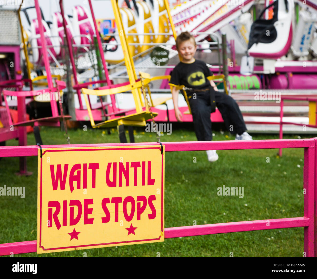 Happy young boy having fun on a fairground ride. Swings and roundabouts. UK. Warning sign Wait Until Ride Stops. Stock Photo