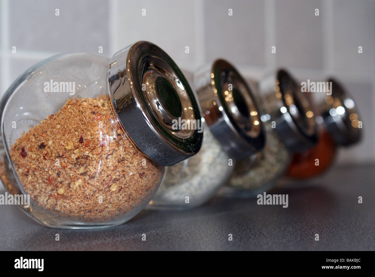 Row of four glass spice jars with chrome lids, containing various herbs