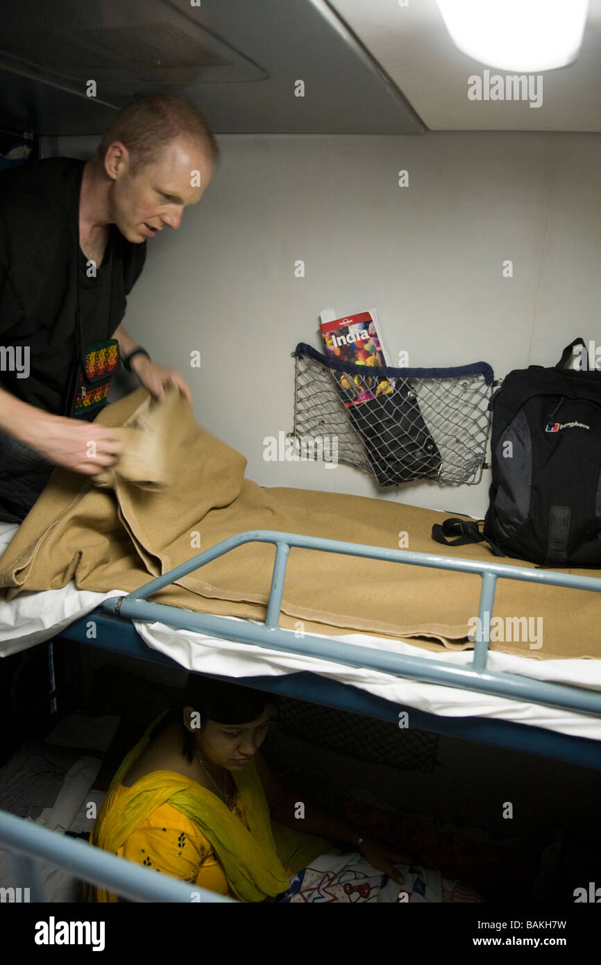 Western European passenger makes his bed in a sleeper compartment of an Indian train. There is an Indian woman passenger below. Stock Photo