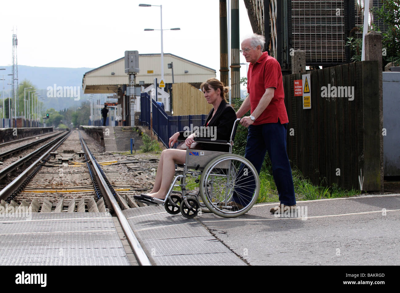 Female invalid wheelchair user and male carer using a railway level crossing England UK Stock