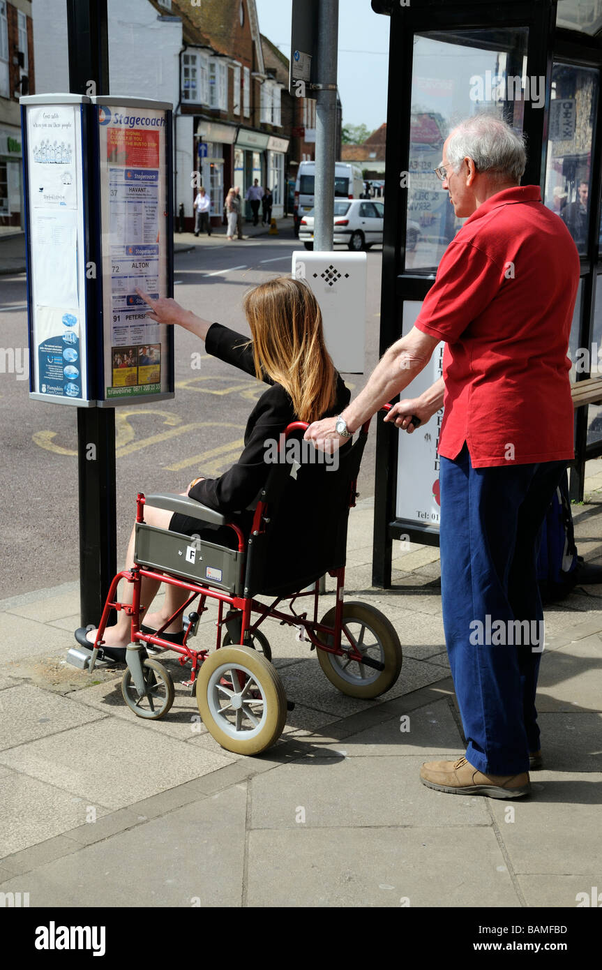 Female invalid wheelchair user and male carer checking bus timetables at a bus stop England UK Stock Photo