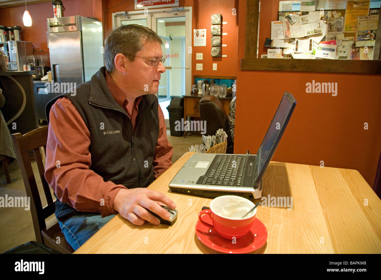 A man works on his lap top computer and drinks a cappuccino coffee at a coffee house in a downtown business area Stock Photo