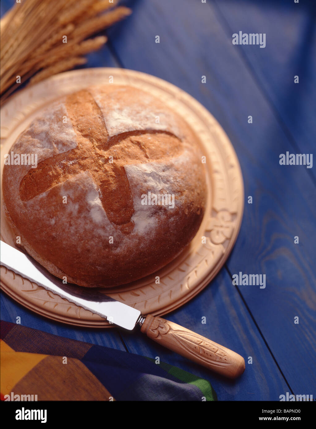 A loaf of round home-style bread sits on a round wooden cutting board with a blue wood background Stock Photo