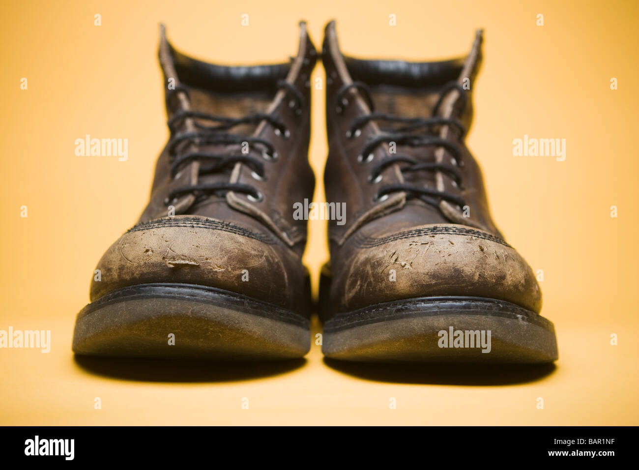 Frontal view of heavy scuffed brown work boots on yellow background. Selective focus on toes, slight vignette lighting Stock Photo