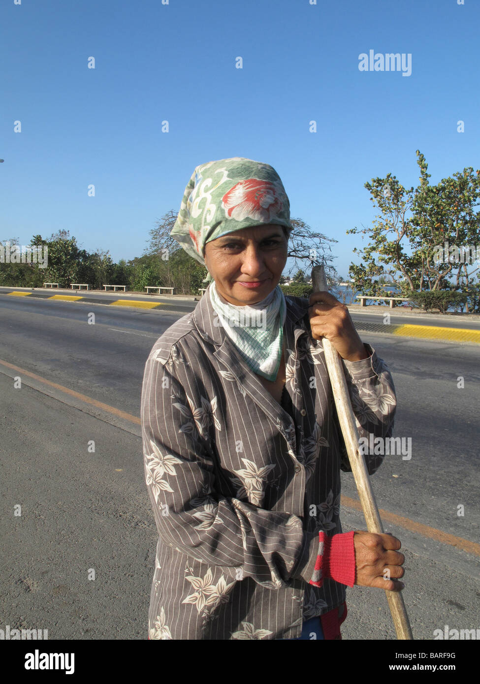 well groomed and dressed, a smiling woman takes a break from sweeping the cycle lane leaving the city of Cienfuegos, Cuba Stock Photo