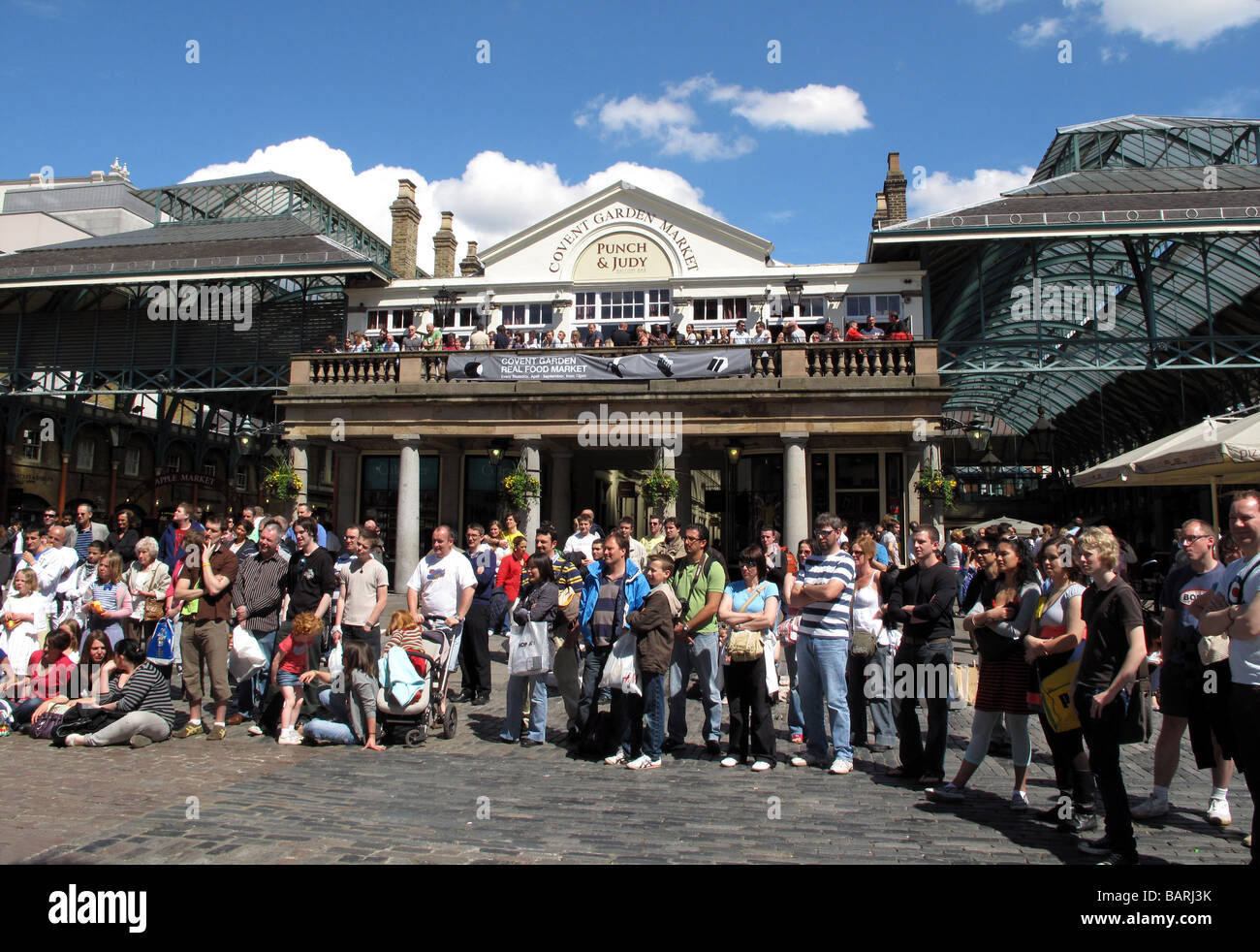 Crowds of tourists watching street artists busking in the sunshine outside Punch and Judy pub at Covent Garden Market in London. Stock Photo