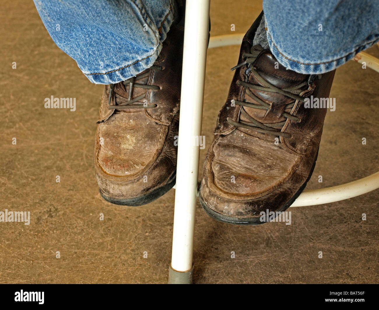 pair of work boots workboots shoes resting on the bottom rung of a stool with bluejeans cuffs visible, brown and worn Stock Photo
