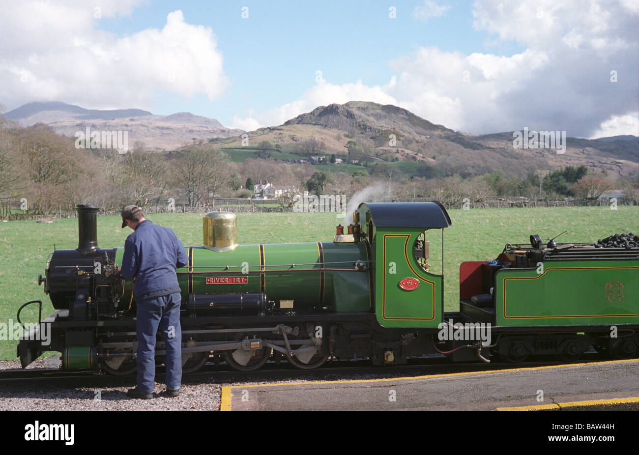 An engineer maintains a 15 inch gauge steam locomotive train before departure with the hills of Eskdale in the background. Stock Photo