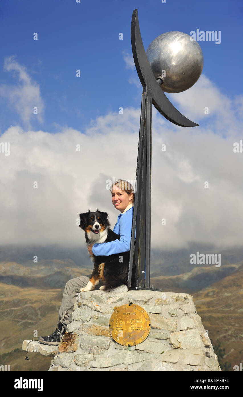 A young woman hiker with her dog sitting on a sculpture of Neptune on the Planets Footpath in the Swiss alps. Stock Photo