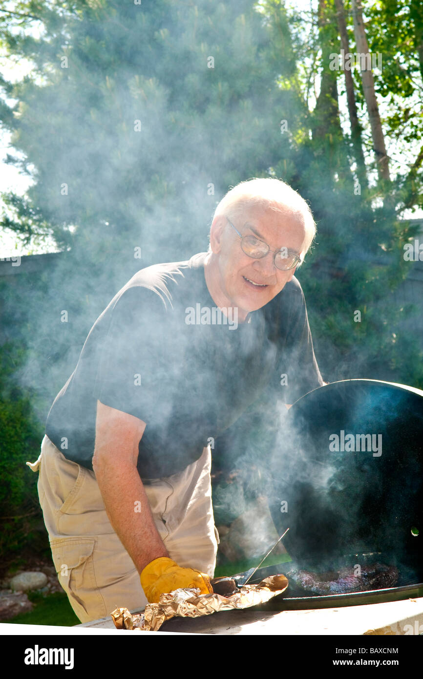Elderly middle age caucasian man cooking meat on a charcoal barbeque with smoke emanating from grill Stock Photo