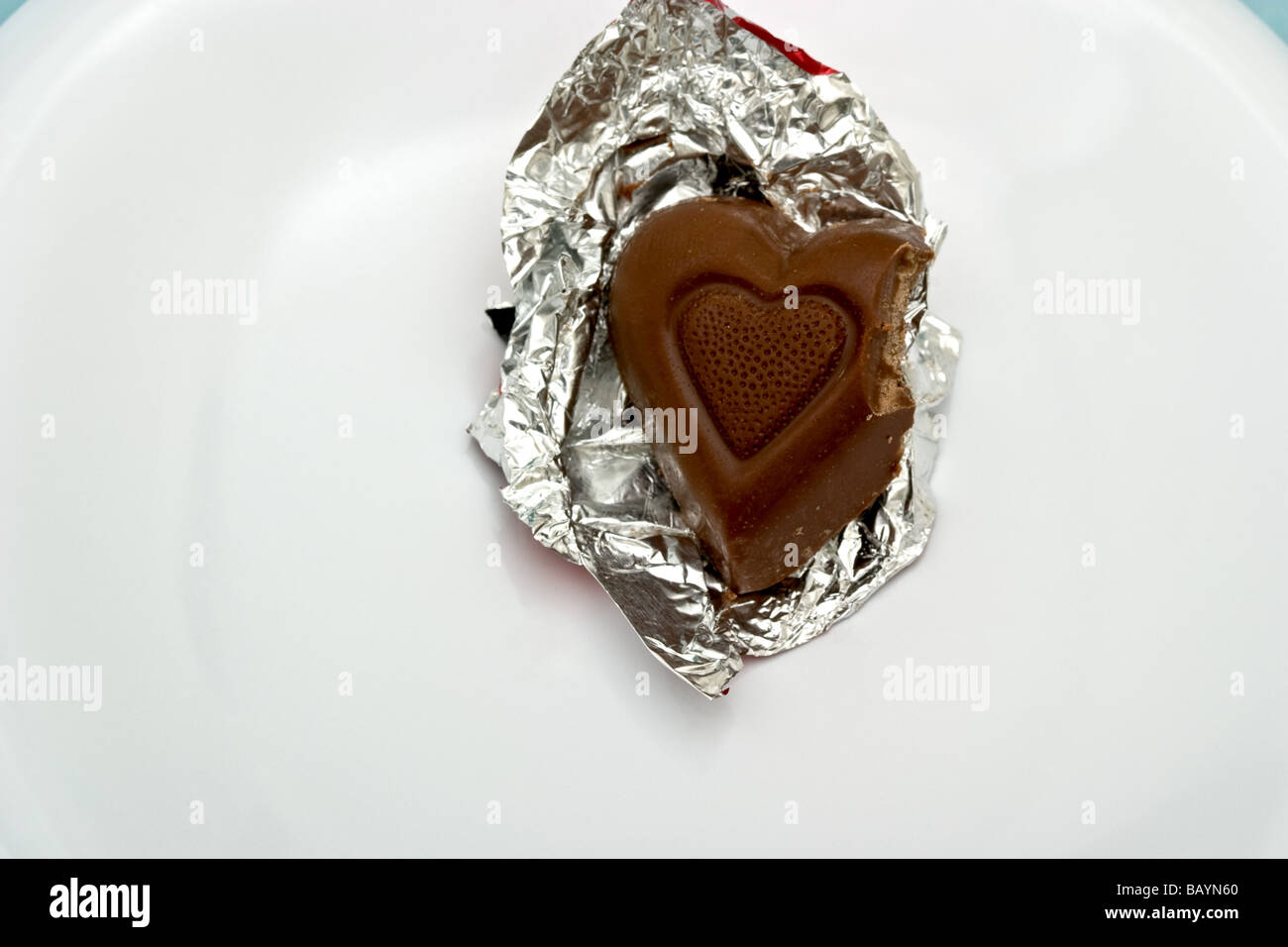 Single heart shaped chocolate candy in aluminum foil wrapping with a bite taken out of the corner on a plate Stock Photo
