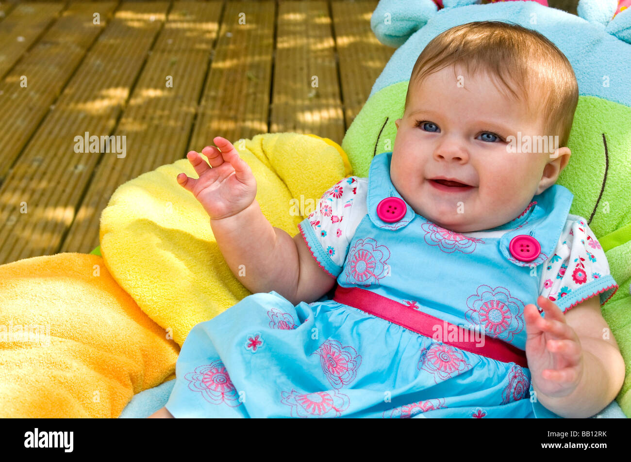 Horizontal close up portrait of a cute six month old baby girl sitting on a colourful cushion in the garden on a sunny day Stock Photo