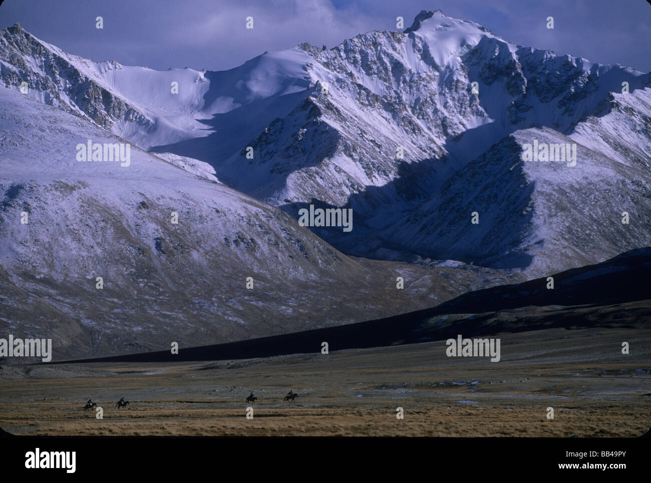 Kyrgyz horsemen ride below snow covered peaks of the Wakhan range, in the Big Pamirs, Wakhan Corridor, Badakshan Stock Photo