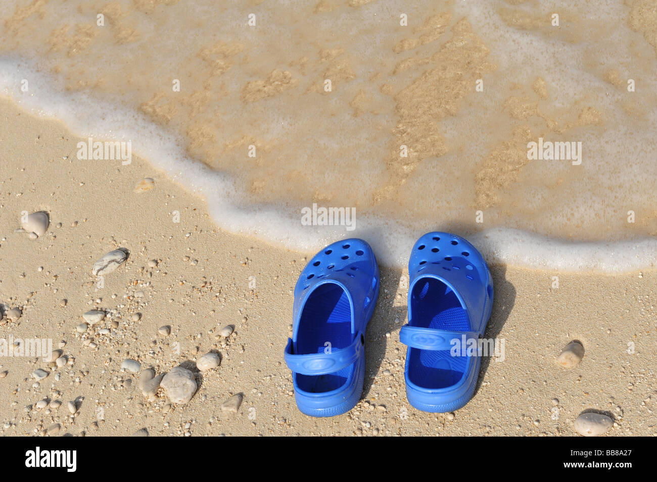 Pair of blue beach shoes or crocs at the edge of the sea with a small wave and sand Stock Photo