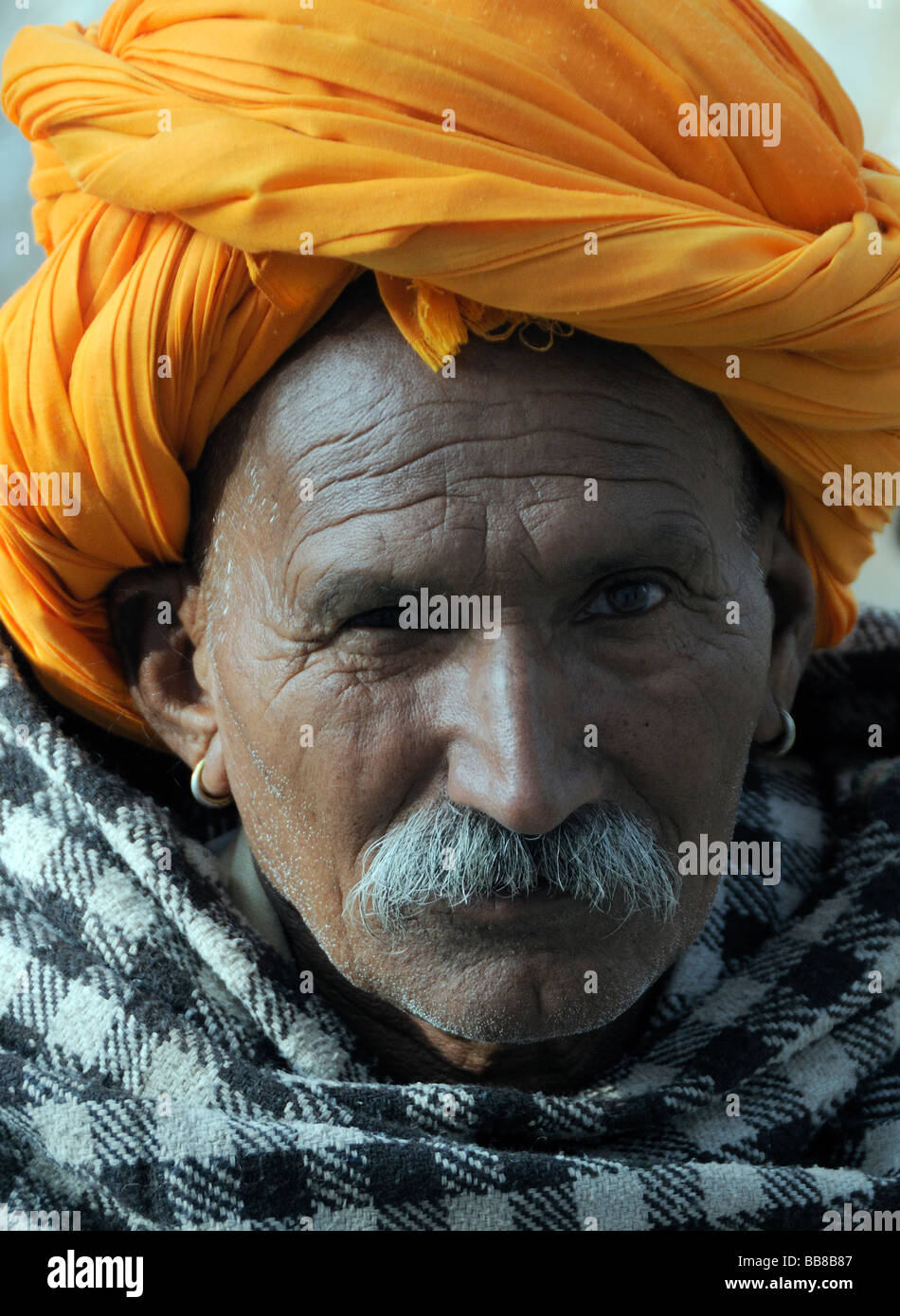 A  man with ear rings, a moustache and an orange turban is wrapped in a chequed wool shawl against the morning chill. Stock Photo