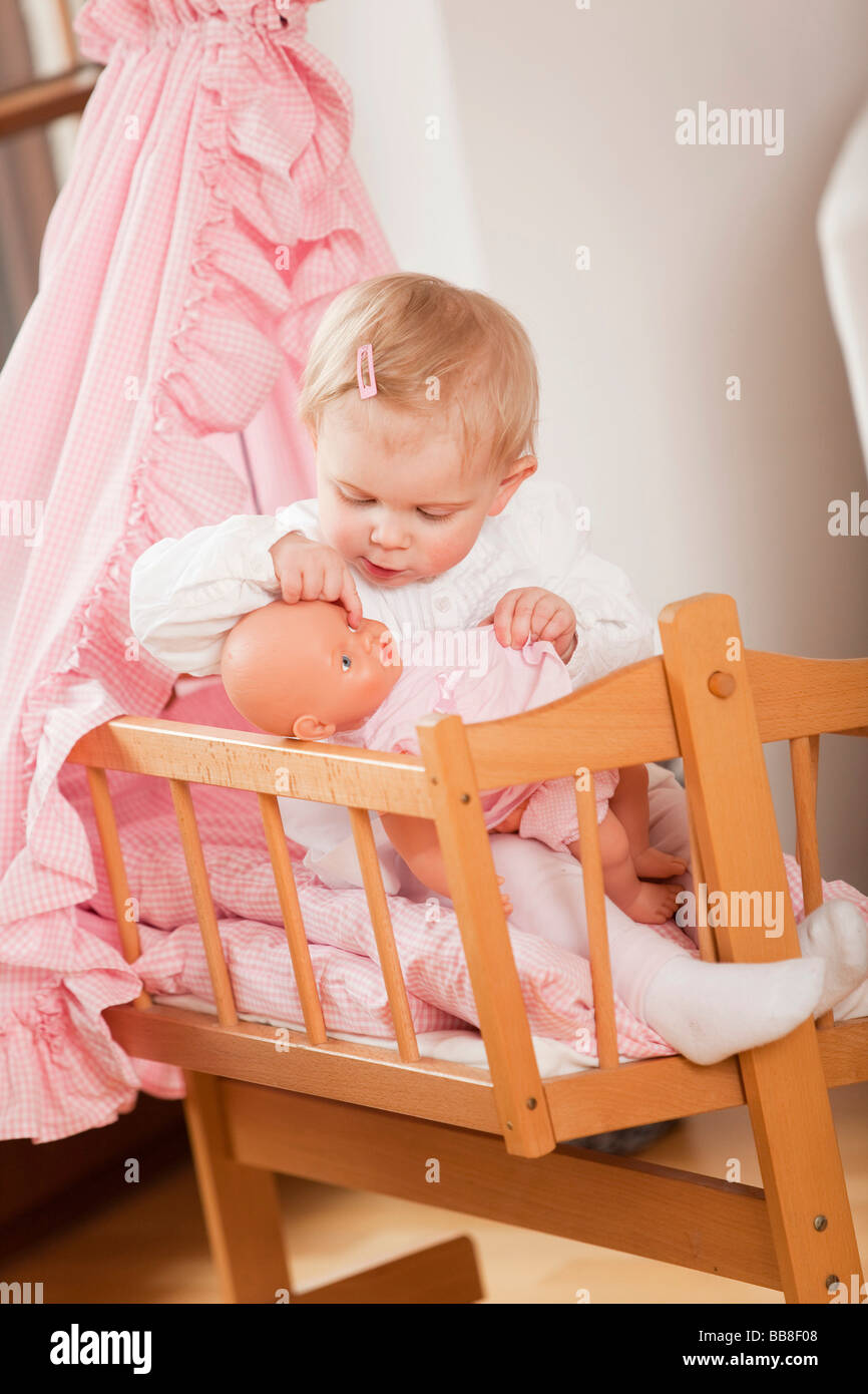 Young girl, 1 year old, sitting in her doll's pram and playing with a doll Stock Photo