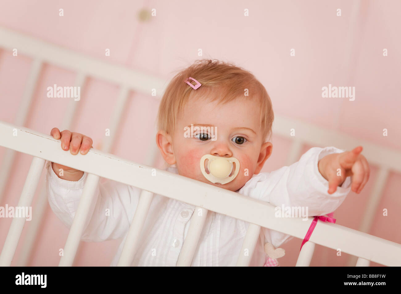 Young girl, 1 year old, with a dummy, looking over the bars of her cot, pointing at something Stock Photo