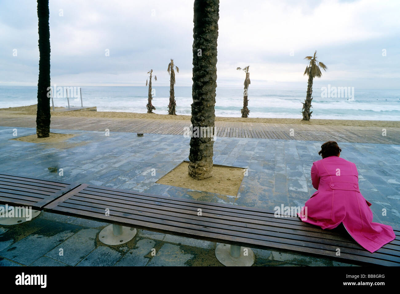 Lonesome woman wearing a pink coat sitting on a bench and looking at the ocean, promenade, Barcelona, Spain, Europe Stock Photo
