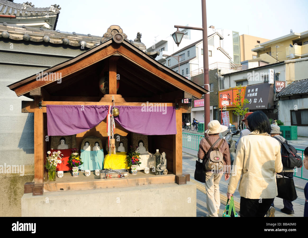 People pass a small shrine-like roadside structure in Ibaraki, a suburb of Osaka, Japan. Such street scenes are very typical. Stock Photo