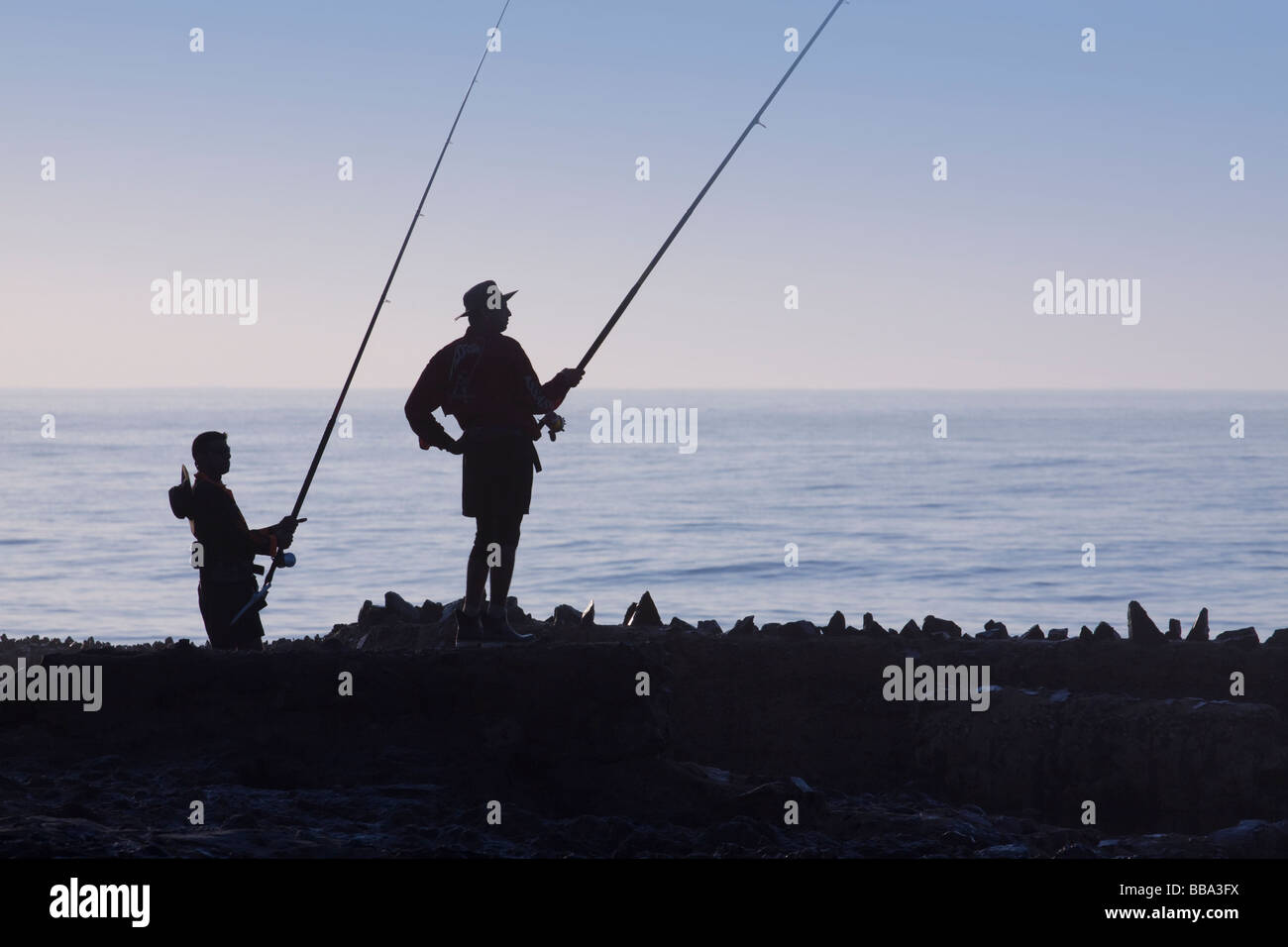 Two fisherman in silhouette at dawn fishing off seaside rocks. Backlit. Silhouette. Durban, KwaZulu Natal, South Africa. Stock Photo