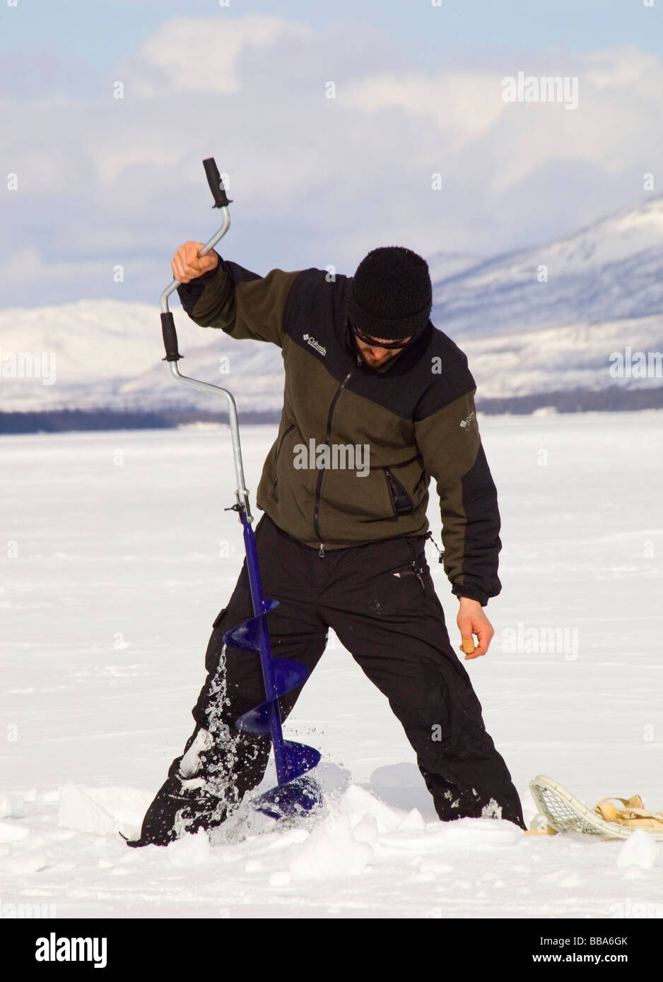 Man ice fishing, drilling a hole with an ice auger, Fox Lake, Yukon