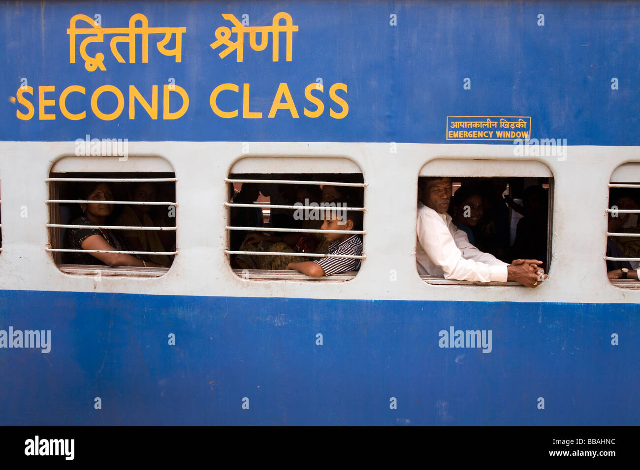 People sit inside of a second class carriage on a train in India. Many of the windows have bars on them. Stock Photo