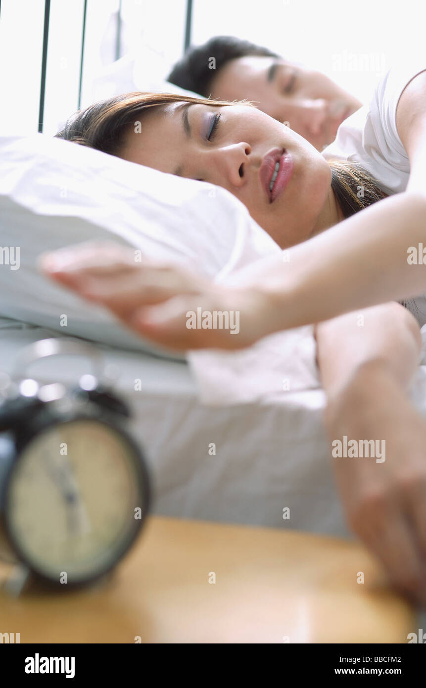 Couple lying on bed, woman reaching to switch of alarm clock in foreground Stock Photo