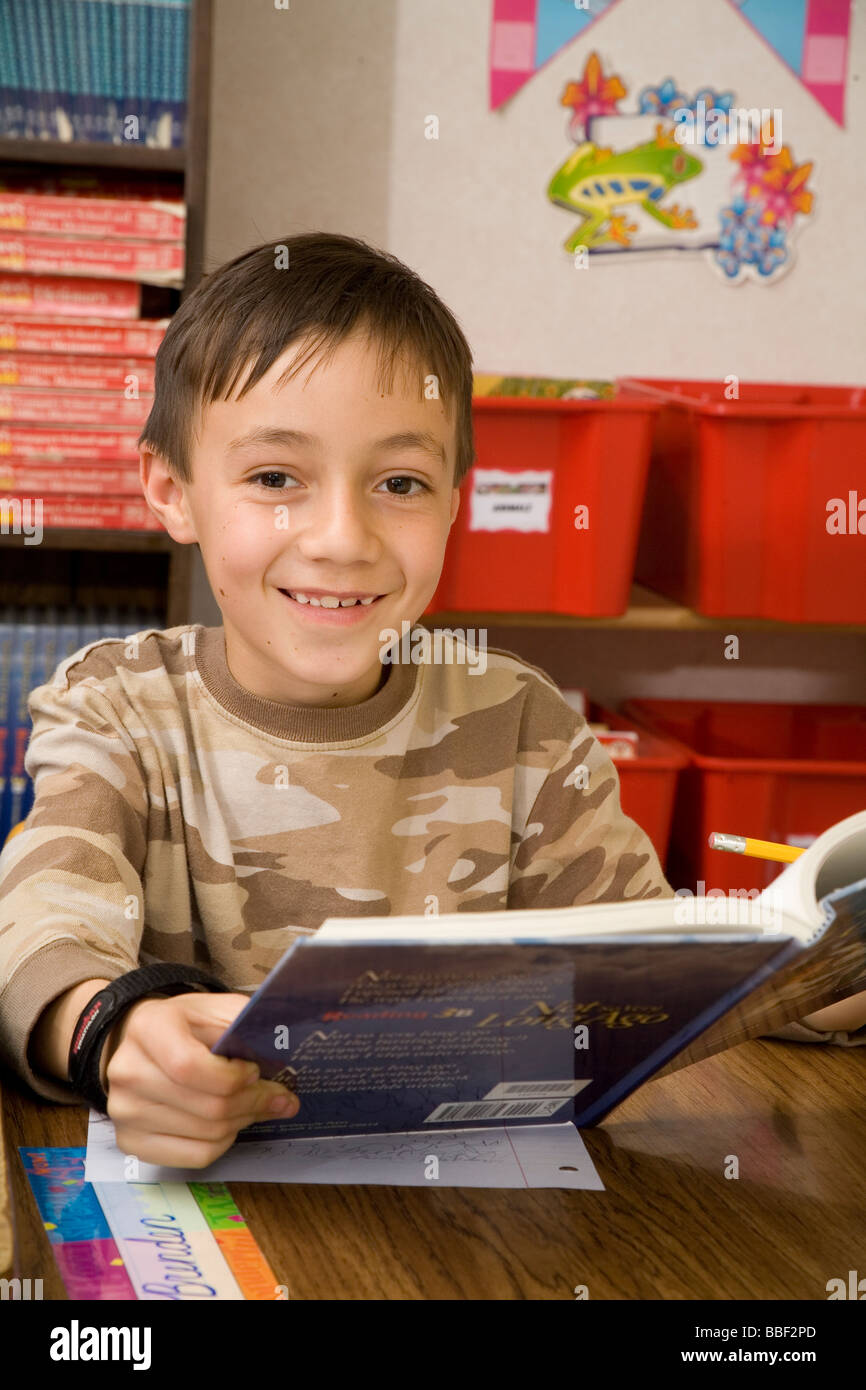 Portrait young person child children boy 8-10 year old reading book in classroom looking smiling camera 3rd grade eye contact United States MR Stock Photo