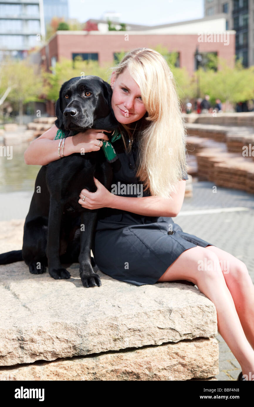 Blond white caucasian girl woman with black labrador pet guide dog in an urban park setting. Stock Photo