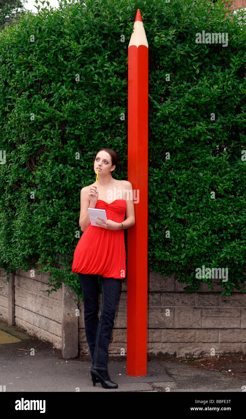 Young woman standing by giant-sized red pencil, with a notepad, waiting for inspiration Stock Photo
