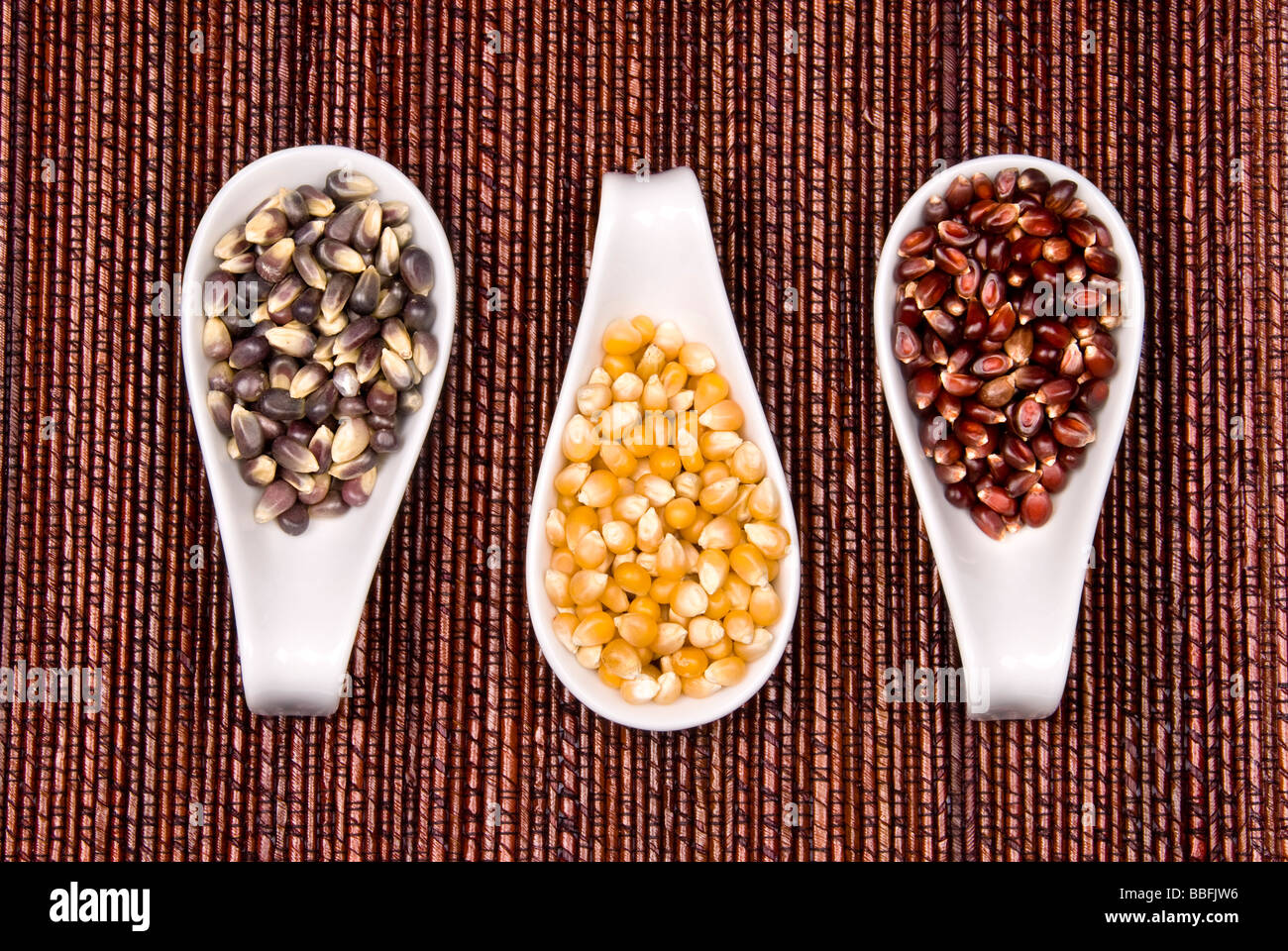 Three types of popcorn including crimson red black Jewell and classic yellow sit in spoonfulls on a bamboo place mat Stock Photo