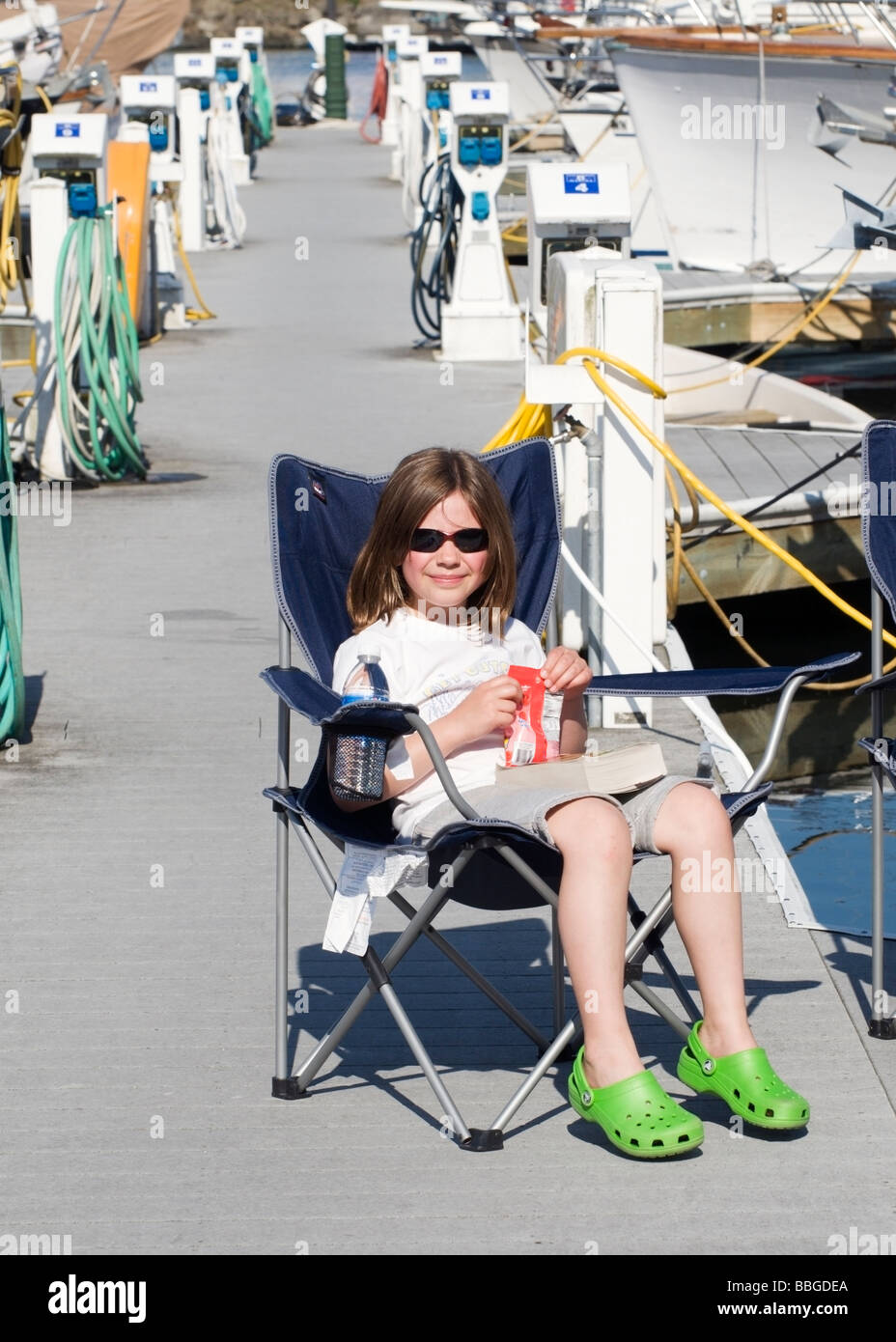 A little girl with book, water, and snack relaxes in a chair on the dock at Foss Waterway Marina in Tacoma, Washington. Stock Photo