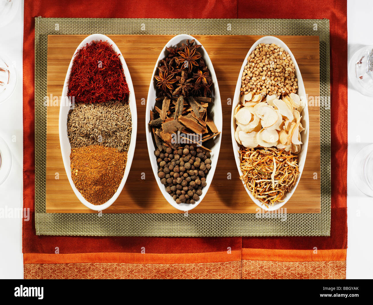 A selection of different spices in three white china bowls on a wooden placemat and red background Stock Photo