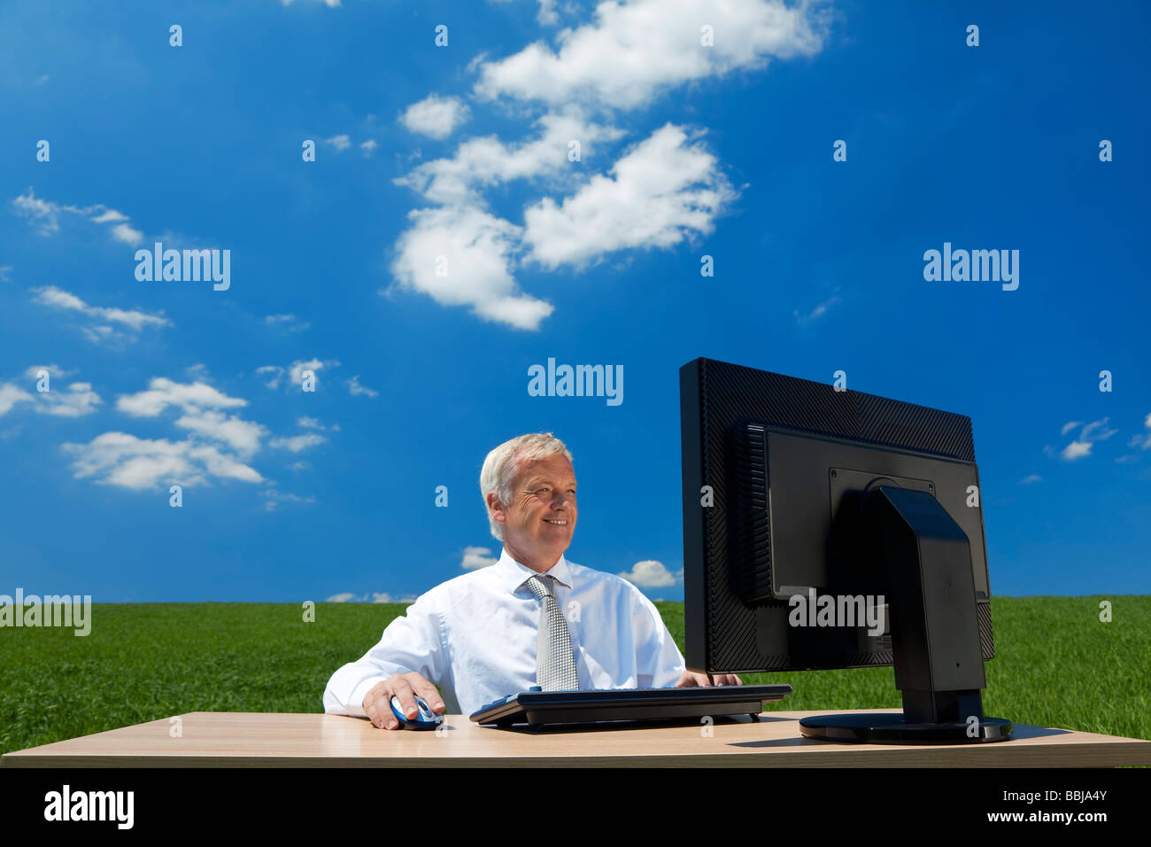 Business concept shot showing an older male executive using a computer in a green field with a blue sky and fluffy white clouds Stock Photo