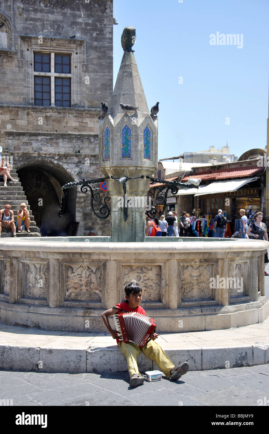 Boy playing accordion by Castellania fountain, Ippokratous Square, Old Town, City of Rhodes, Rhodes, Dodecanese, Greece Stock Photo