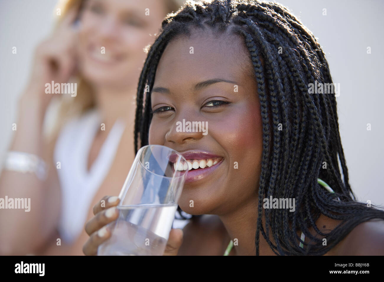 Portrait of a young african woman with waterglass outdoors smiling at the camera, a female friend in the background out of focus Stock Photo