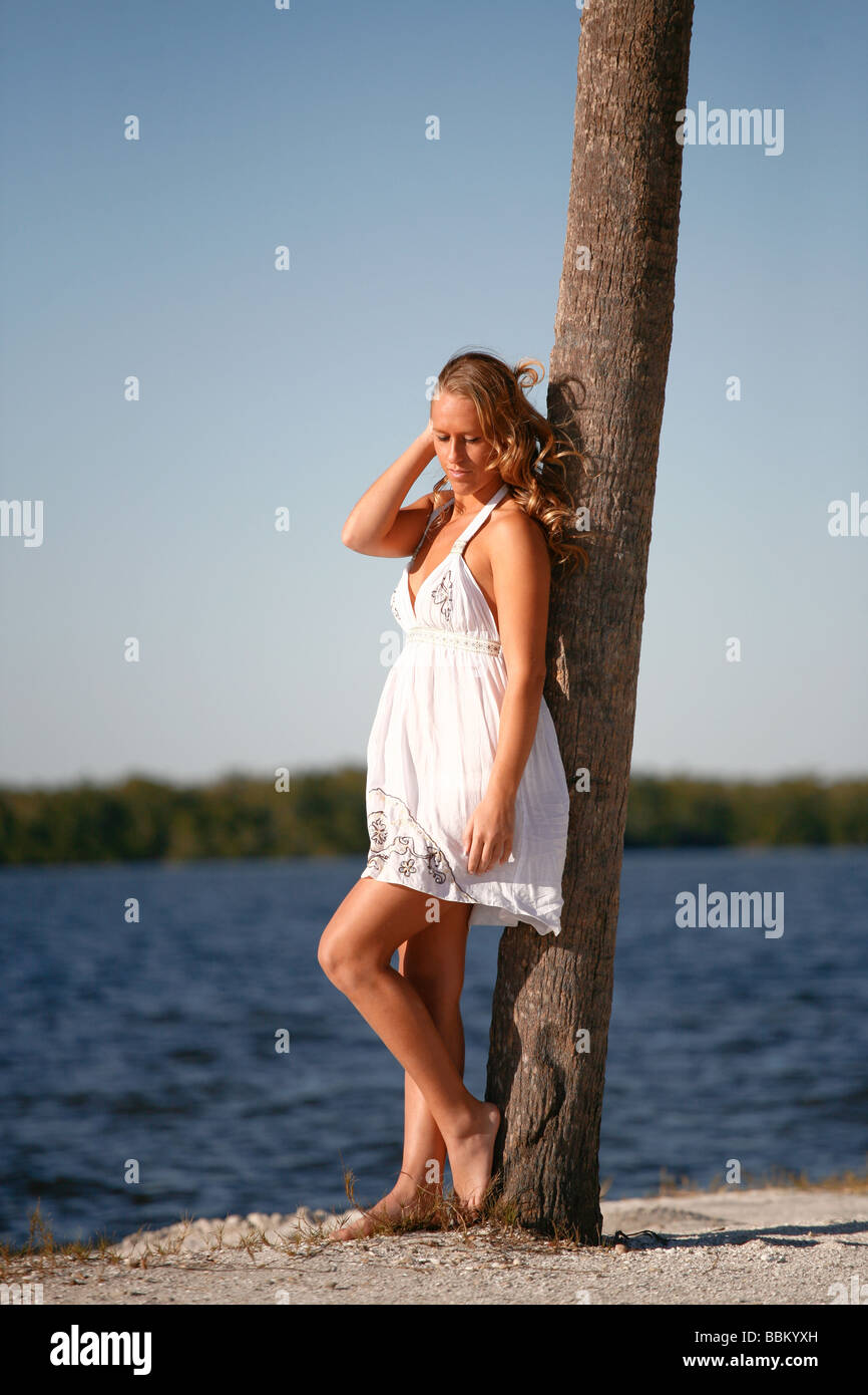 Young white woman leaning on palm tree on island beach in white sundress, barefoot. Stock Photo