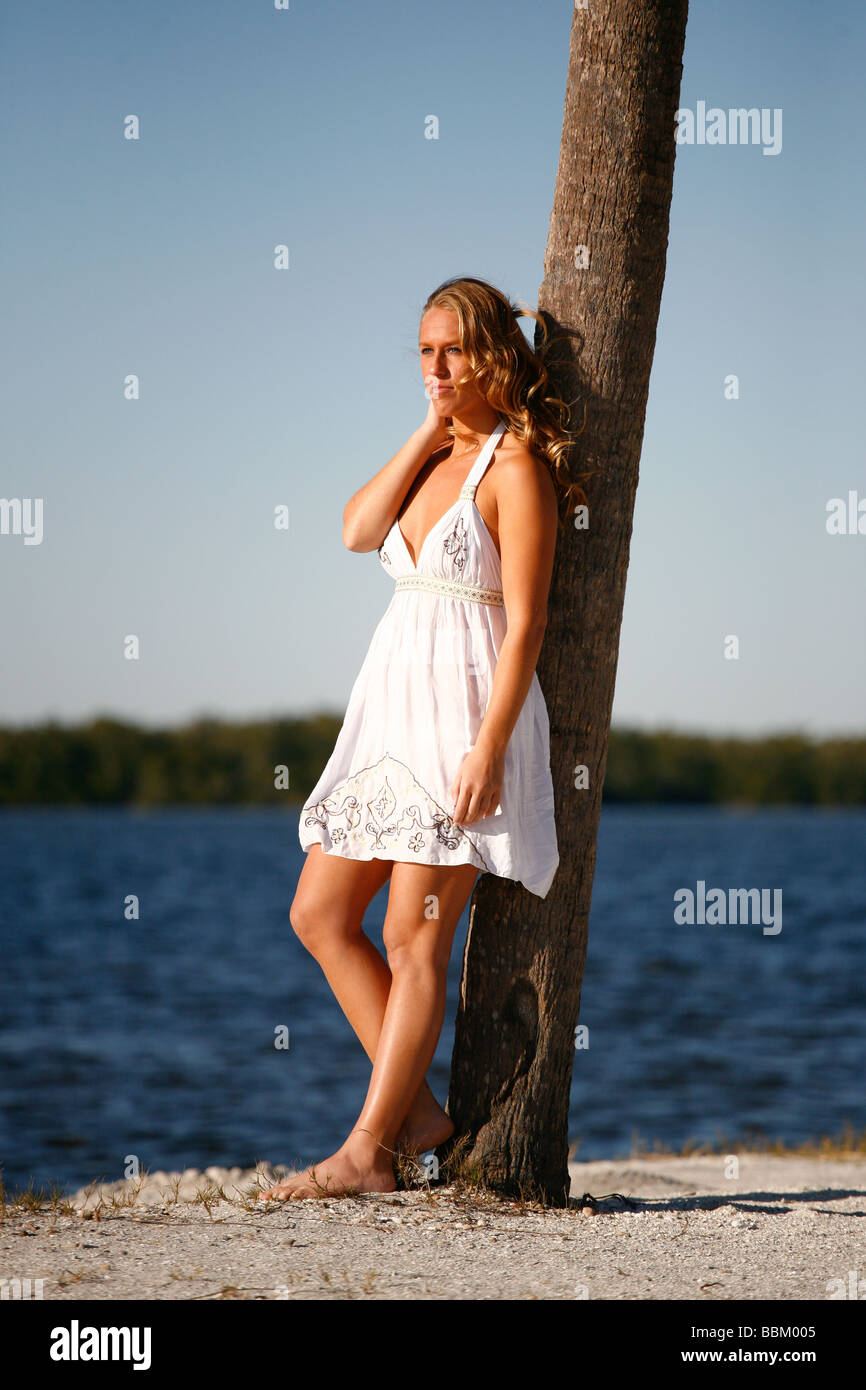 Young white woman leaning on palm tree on island beach in white sundress, barefoot. Stock Photo